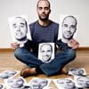 A man is sitting cross-legged on the ground surrounded by scattered black and white print-outs of his face with various expressions.